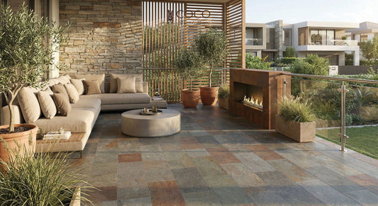 A luxurious outdoor terrace featuring the Sunset Terrace tile look. The floor is laid with Arcadia Series Valley Crest 225x453mm textured porcelain tiles, showing a multi-toned volcanic stone effect in slate grey and warm rust. The space includes a modern corten steel outdoor fireplace, a large beige linen corner sofa, a stacked stone feature wall, timber slatted privacy screens, and potted olive trees, overlooking a landscaped garden.