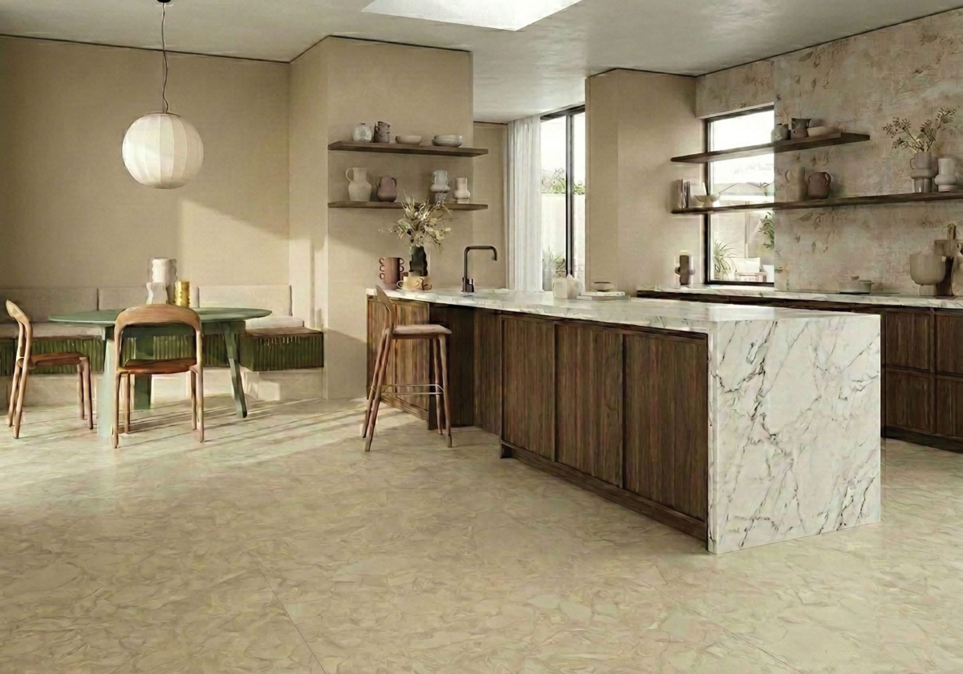 A warm, organic-modern kitchen featuring the Trattoria tile look. The floor is laid with Fragmenta Series Cappuccino 600x600mm creamy beige terrazzo-look tiles. The kitchen wall behind open timber shelving features Babylon Series Villa B 500x1200mm ceramic tiles, displaying a subtle botanical relief pattern with an aged plaster effect. The space includes a marble waterfall island, dark walnut cabinetry, a round green dining table, and a paper lantern pendant light.