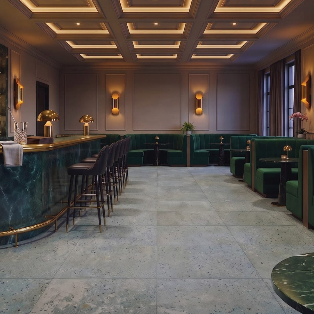Luxury hotel bar with Patina Grigio Azzurro Matt 604x604 blue-grey terrazzo-look porcelain floor tiles, emerald velvet seating, brass lighting and dark stone tables.